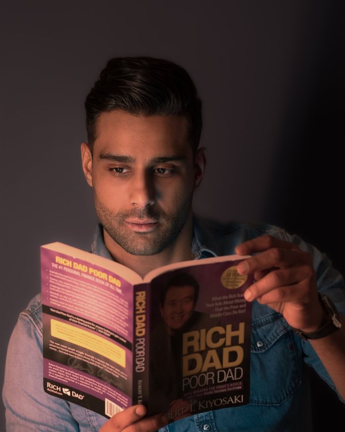 Portrait of a young man intently reading 'Rich Dad Poor Dad' against a gray wall.