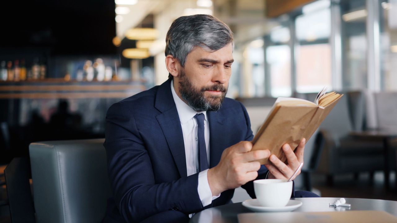Mature man in a suit reading a book while enjoying coffee in a stylish café.