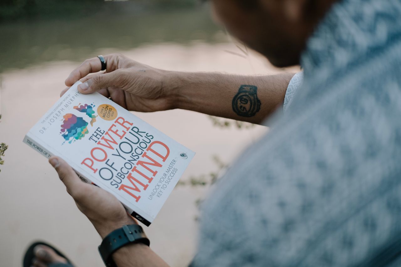 A man reads 'The Power of Your Subconscious Mind' by a riverside, showcasing relaxation and reflection.