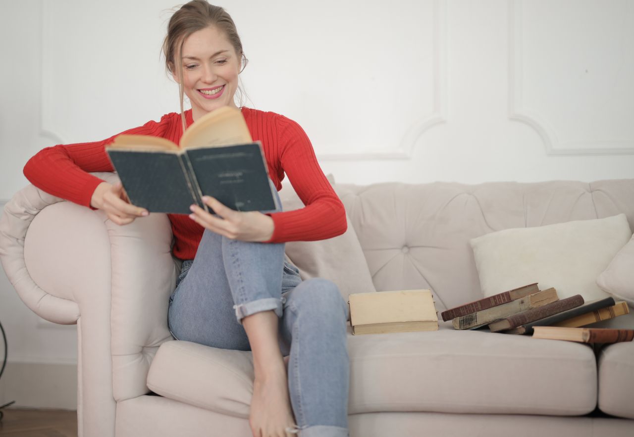 Smiling woman in red sweater reads a book on a cozy sofa, surrounded by vintage books.