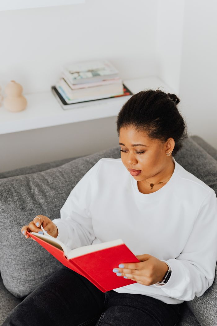 A woman in a white sweater reading a red book while sitting on a gray couch indoors.
