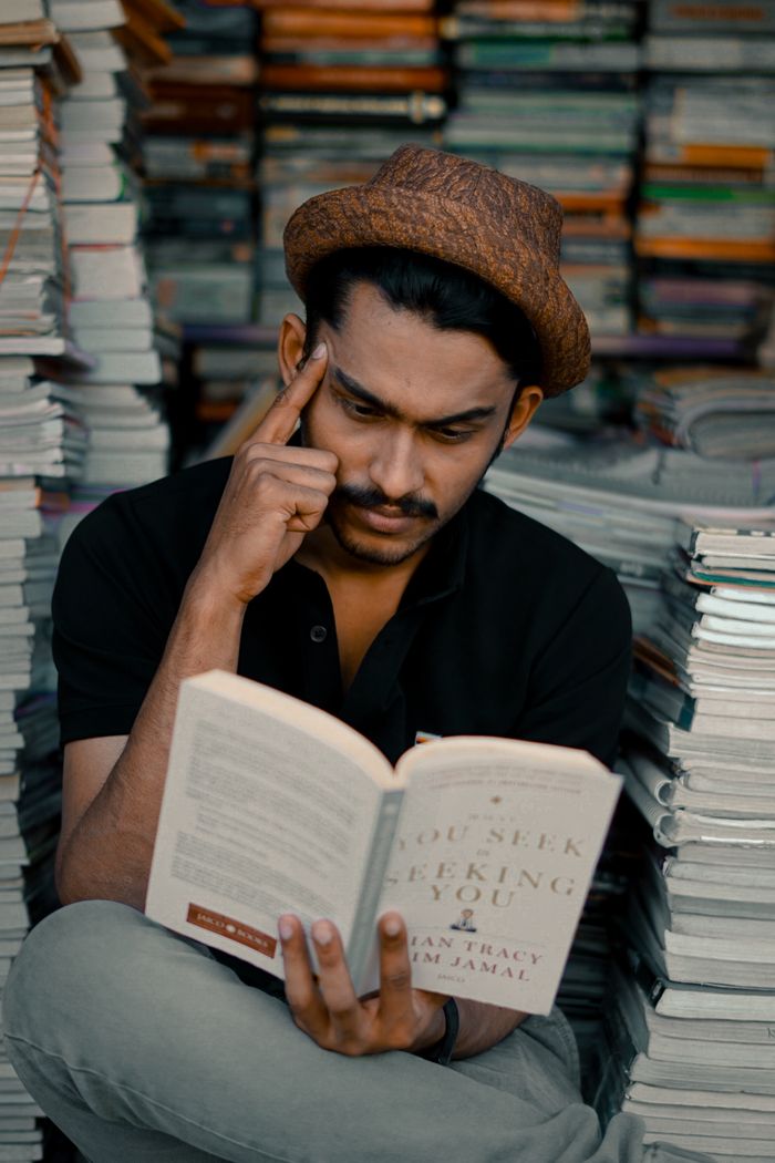A thoughtful man in a hat reads a book in a bookstore surrounded by stacks of books.